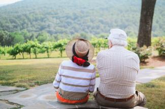 An elderly man and woman sitting together. They have their backs to the camera and are looking out at a vineyard.