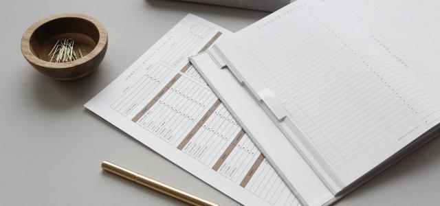 A desk with office supplies on it. In the middle of the desk is an open journal with a heading that reads "Finance Tracker". Under the journal is a piece of paper that reads "Monthly Budget Overview", and has several columns on it for tracking expenses.